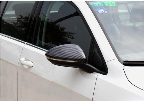 A white car features a carbon-fiber side mirror reflecting nearby trees, parked in an outdoor setting. Stickers, including a 2016 sticker, are displayed on the windshield.