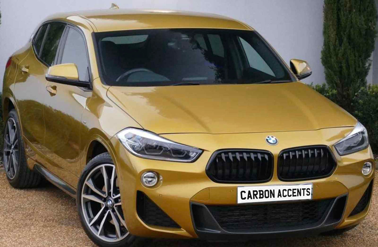A gold BMW parked on a gravel driveway, with a white wall and greenery in the background. The license plate reads "CARBON ACCENTS."