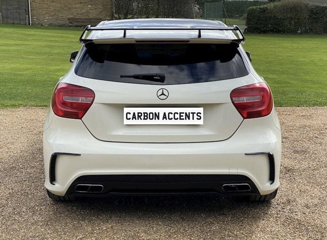 A white car with a spoiler displays a "CARBON ACCENTS" license plate. It's parked on a gravel path, surrounded by a grassy area with hedges and a distant building.