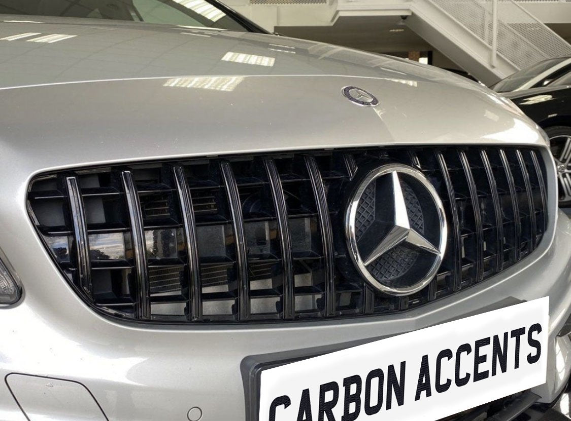 The silver car grille displays a prominent three-pointed logo, surrounded by vertical black slats in a showroom. Below, a white license plate reads "CARBON ACCENTS."