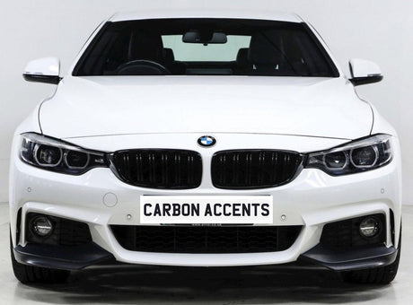 A white BMW car is parked in a studio setting, displaying a front grille and headlights. The license plate reads "CARBON ACCENTS."