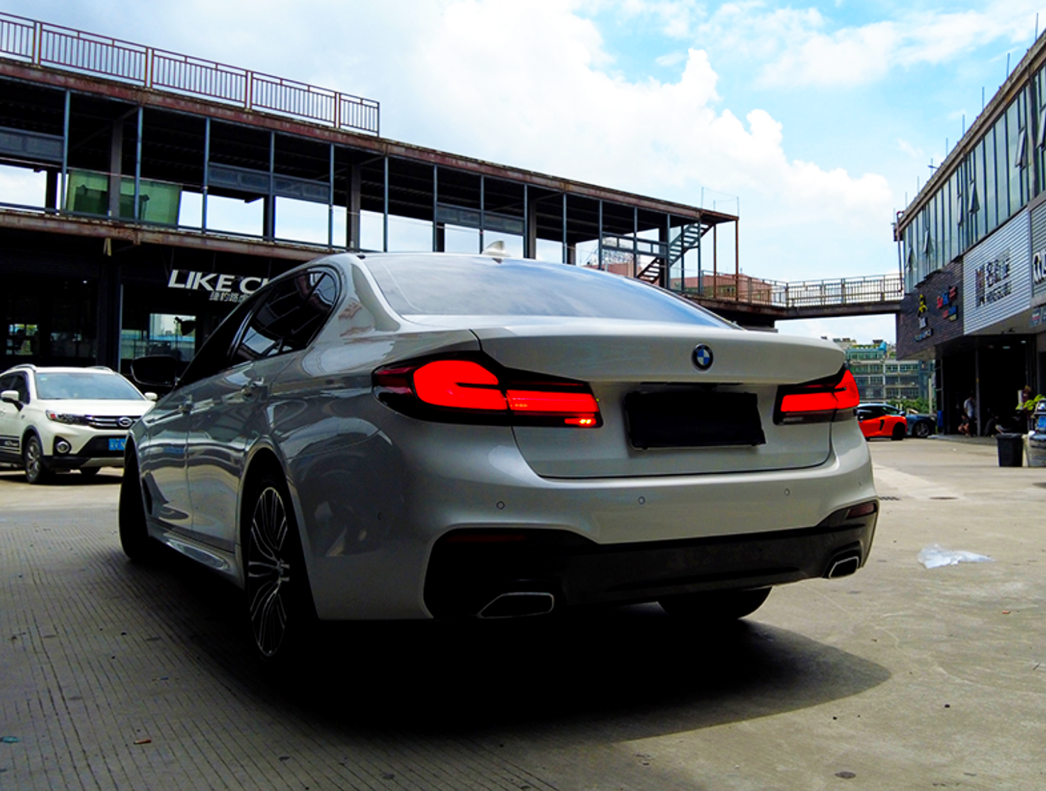 A white BMW car is parked on a concrete surface with its taillights on, surrounded by a building complex. Another car and signs, including "LIKE C," are visible nearby.