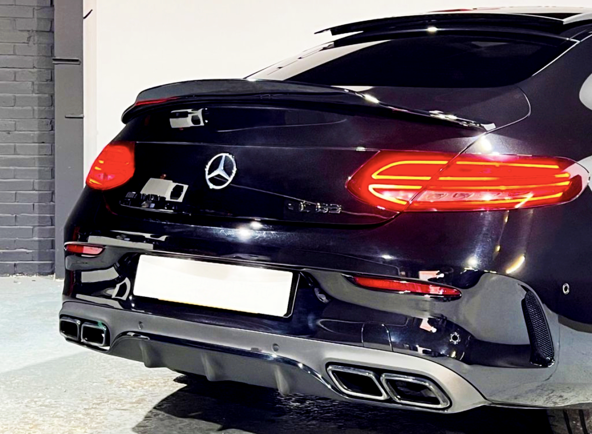 A black car parked indoors, displaying illuminated taillights. The Mercedes-Benz emblem is centered on the trunk. The garage background features grey and white walls.