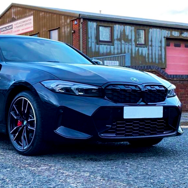 A sleek black car is parked on a gravel surface, with a warehouse featuring rustic metal cladding in the background. A sign on the building reads: "CHAPEL PLANT - SELF ACCOMMODATION & TRANSPORT SERVICES."