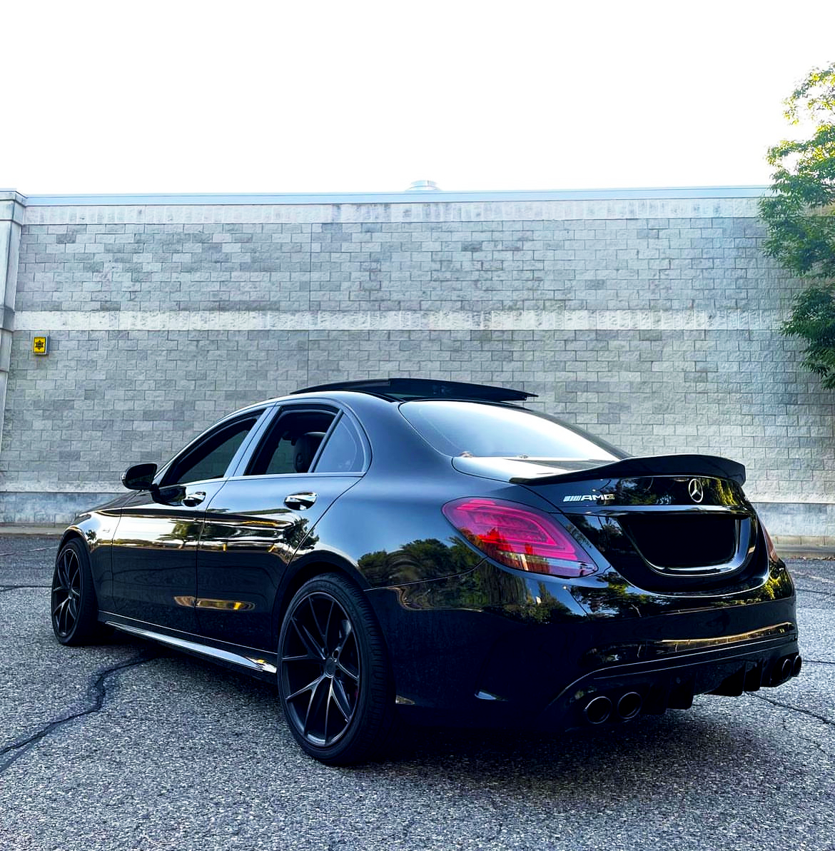A sleek black Mercedes-Benz sedan is parked, featuring the AMG logo. It's set against a light stone wall on an asphalt surface, near trees.