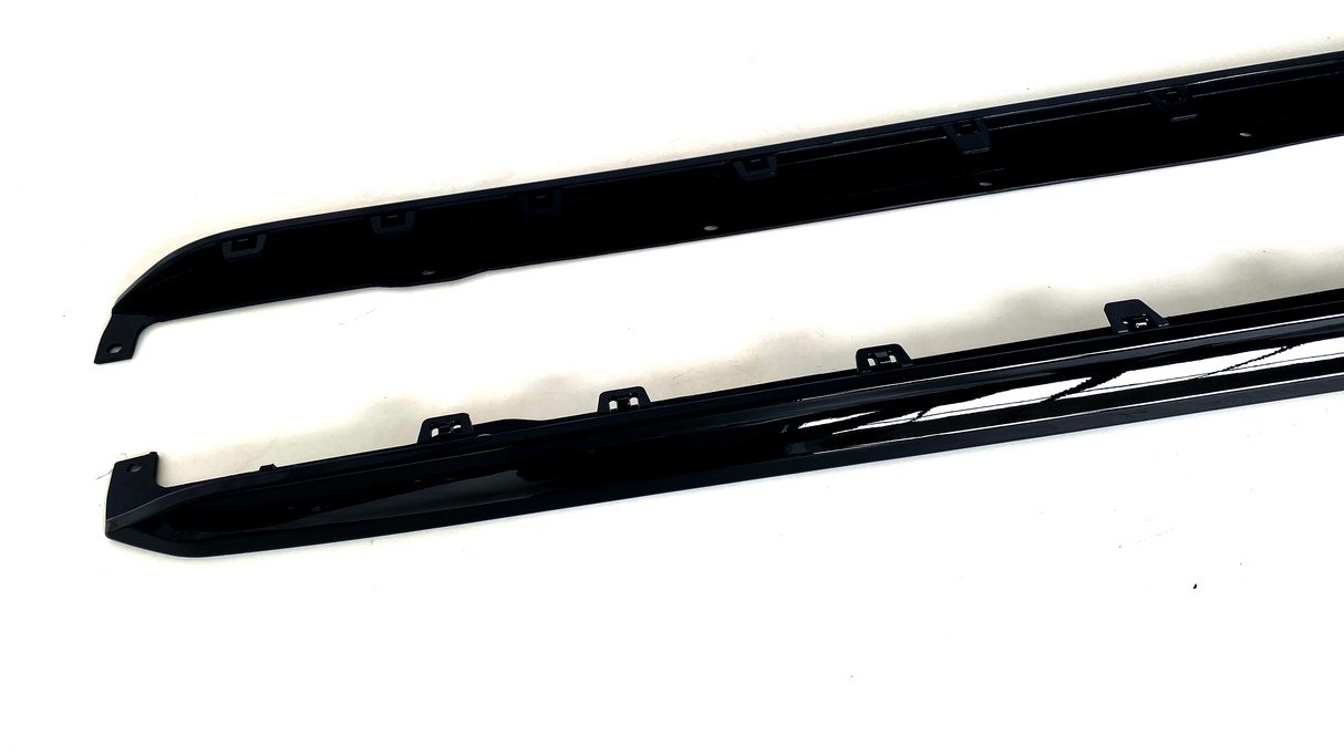 Two black car side skirts are resting on a white surface, featuring several mounting brackets along their length for attachment.