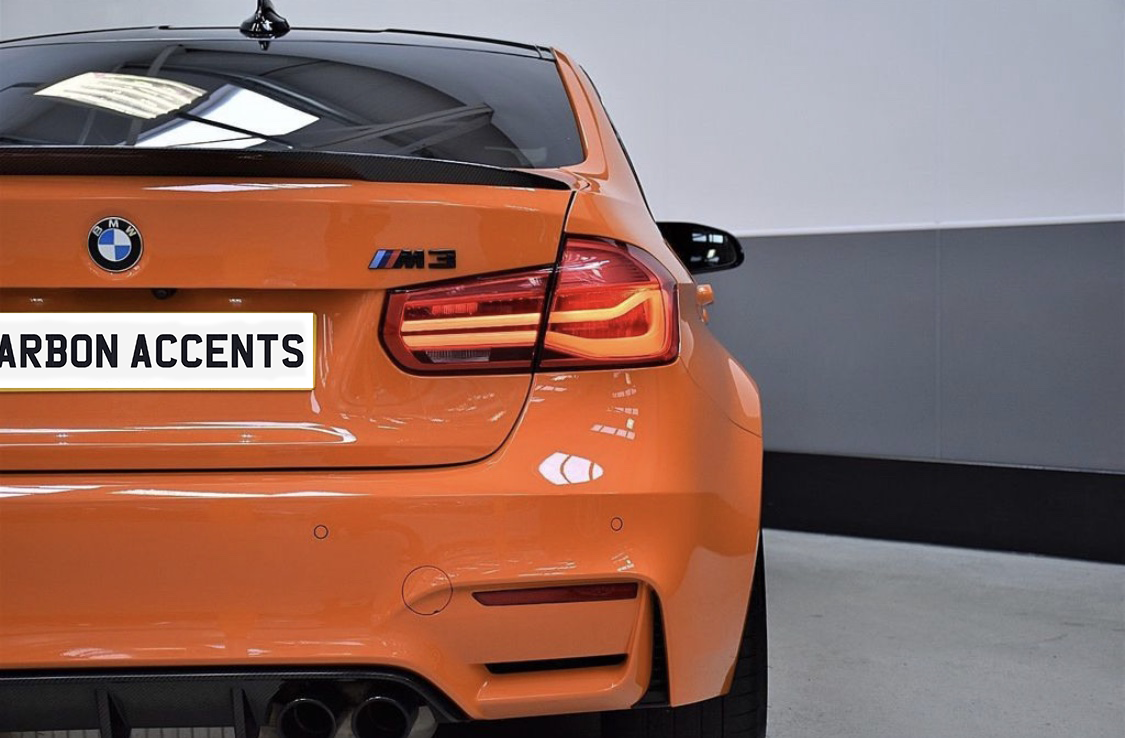 An orange BMW M3 is parked indoors, highlighting its rear view with illuminated taillights. The license plate reads "CARBON ACCENTS," and the environment features a grey wall and concrete floor.