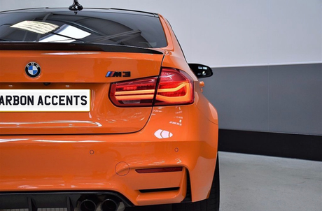 An orange BMW M3 is parked indoors, highlighting its rear view with illuminated taillights. The license plate reads "CARBON ACCENTS," and the environment features a grey wall and concrete floor.