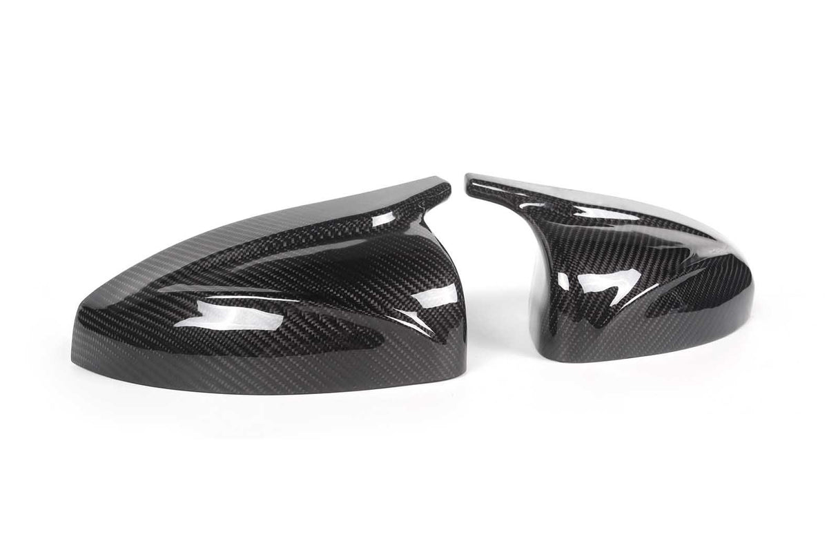 Two sleek, black carbon fiber car mirror covers rest on a plain white surface, reflecting light and showcasing their glossy, patterned finish.