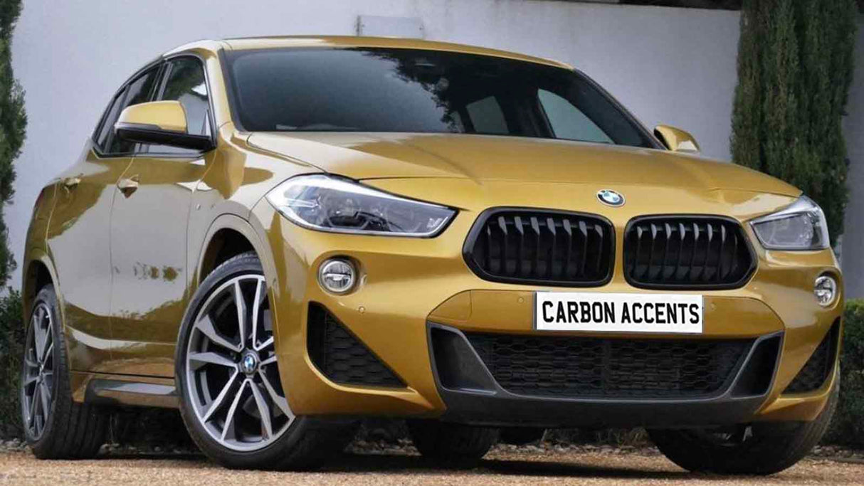 Golden SUV parked on gravel, featuring prominent black grill and sleek design. License plate reads "CARBON ACCENTS." Surrounded by tall shrubs against a white wall backdrop.