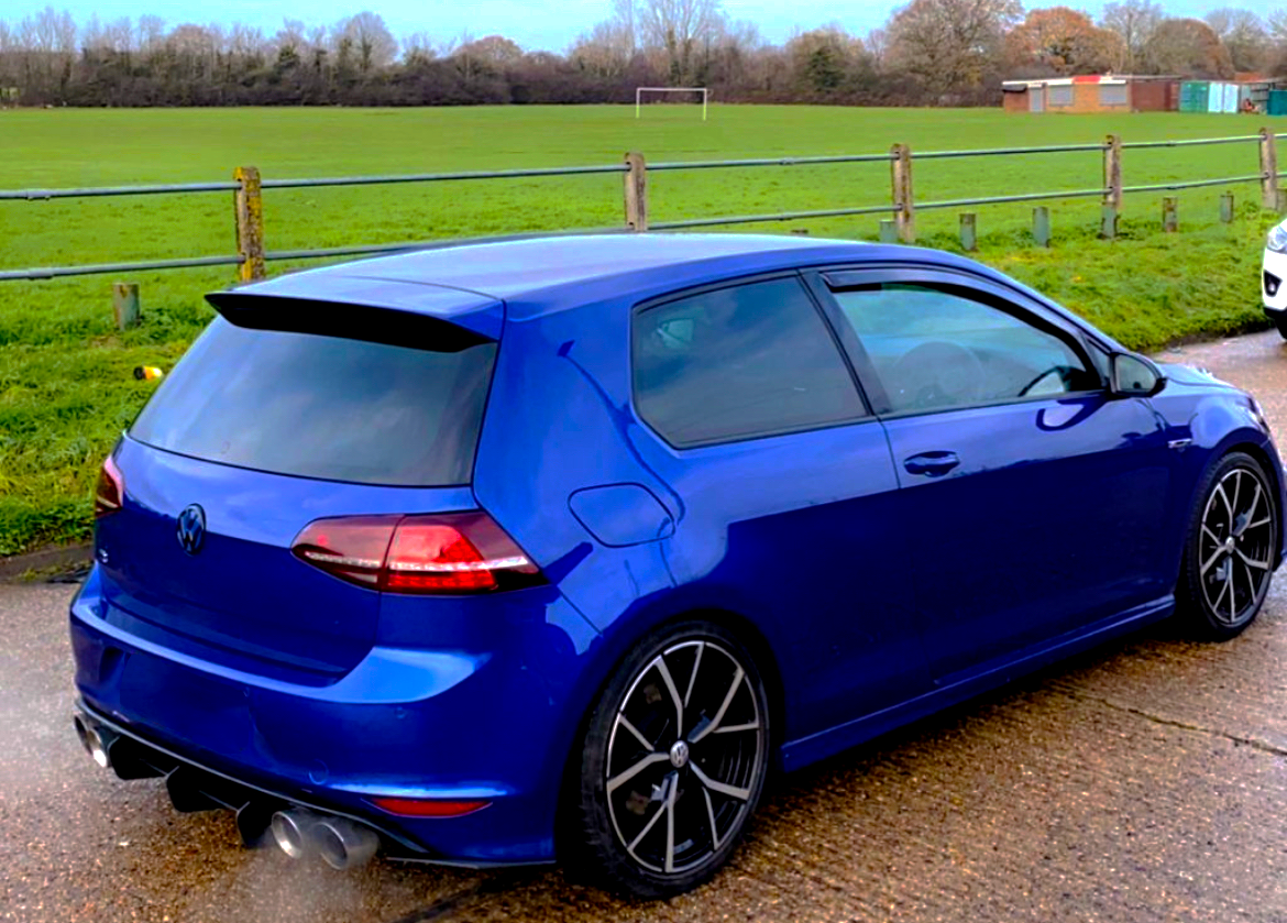 A blue car is parked, emitting exhaust, beside a grassy field with a soccer goal and wooden fencing.