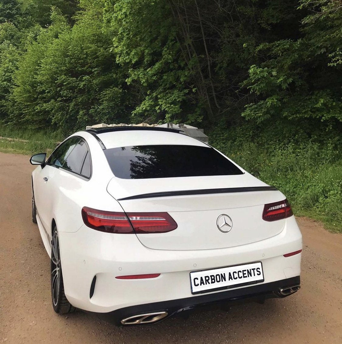 A white Mercedes-Benz coupe is parked on a dirt road, surrounded by lush green trees. The license plate reads "CARBON ACCENTS."