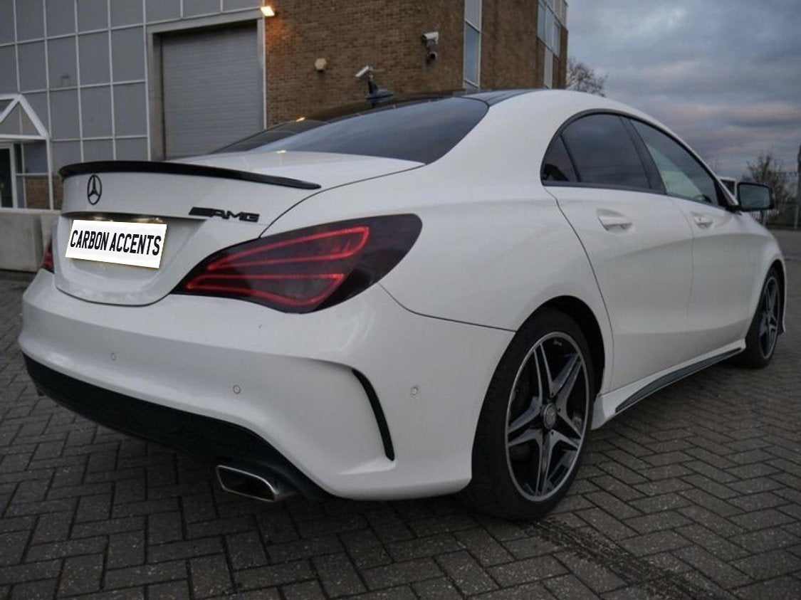A white Mercedes-Benz AMG sedan is parked on a brick-paved area beside a building. The license plate reads "CARBON ACCENTS." The environment is overcast.