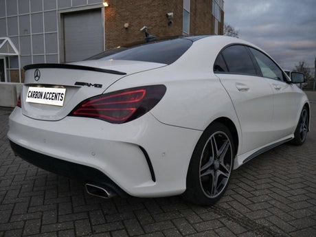 A white Mercedes-Benz AMG sedan is parked on a brick-paved area beside a building. The license plate reads "CARBON ACCENTS." The environment is overcast.
