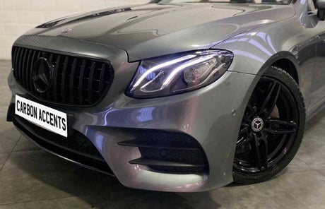 A silver car with illuminated headlights is parked indoors. A prominent grille features a star emblem. The license plate reads "CARBON ACCENTS." Black alloy wheels enhance the sleek design.