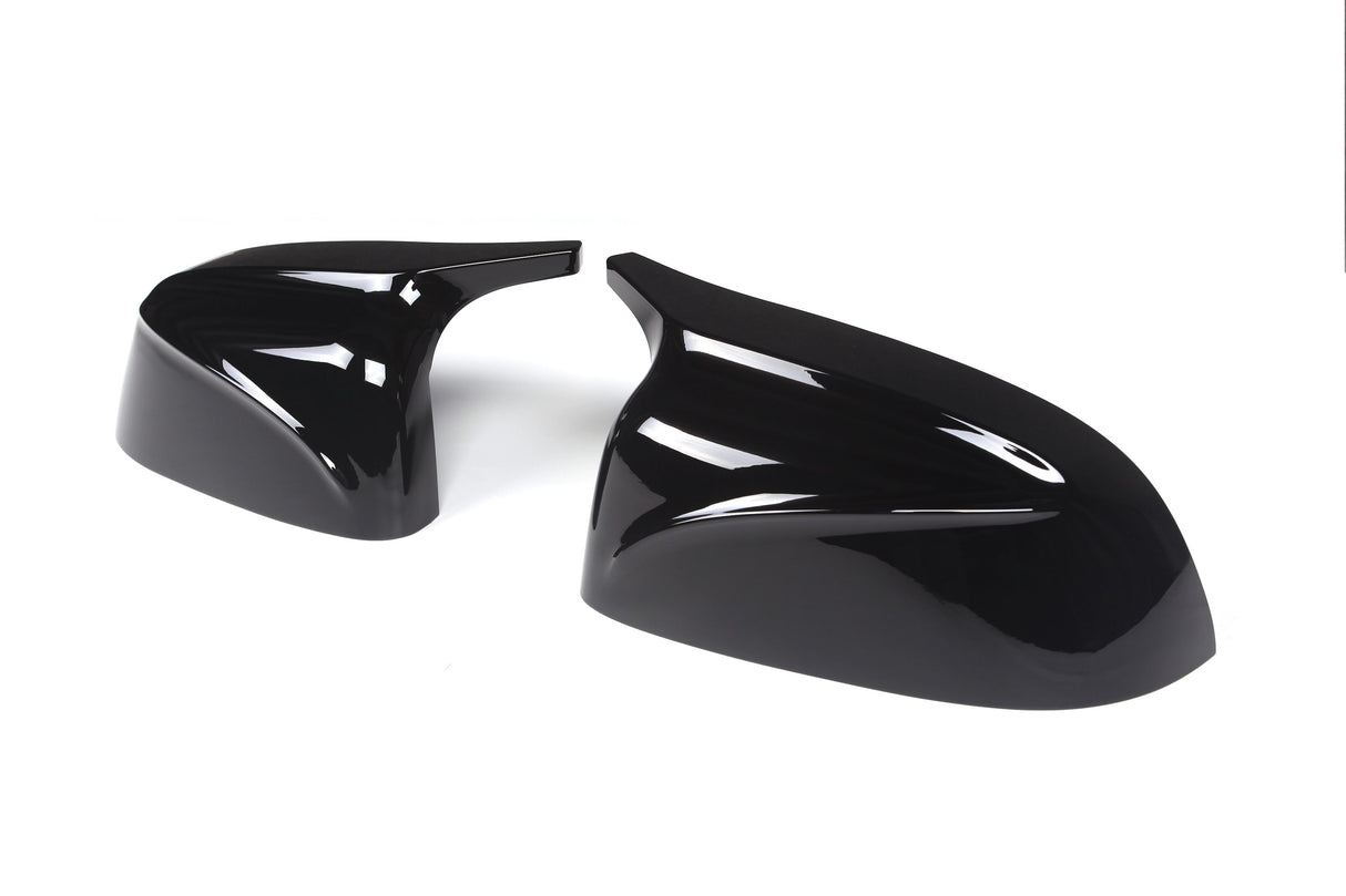 Two glossy black car mirror covers are lying on a plain white background. The covers have sleek aerodynamic designs, reflecting light prominently on their curved surfaces.