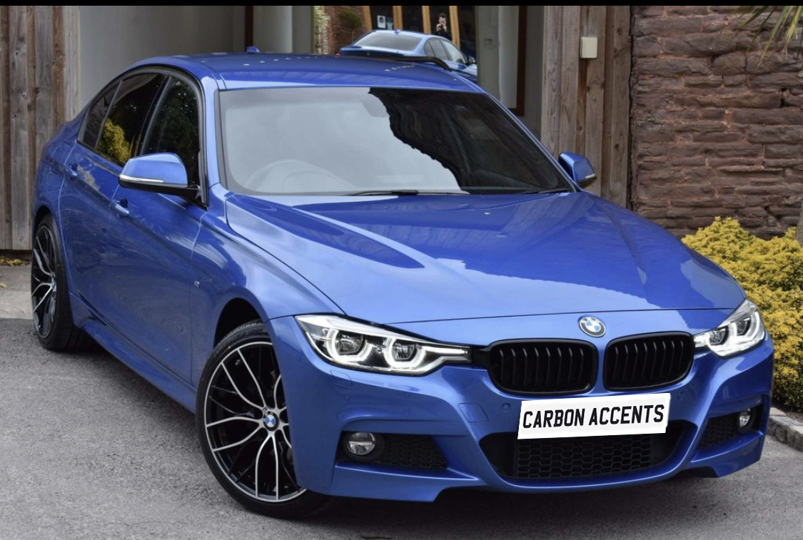 Blue BMW car parked outside a wooden building, featuring black rims and a license plate that reads "CARBON ACCENTS," with bushes and rock wall in the background.