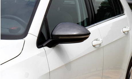 A car's side mirror with a carbon fiber finish is attached to the side of a white vehicle. The door handle and window are visible in an outdoor setting.