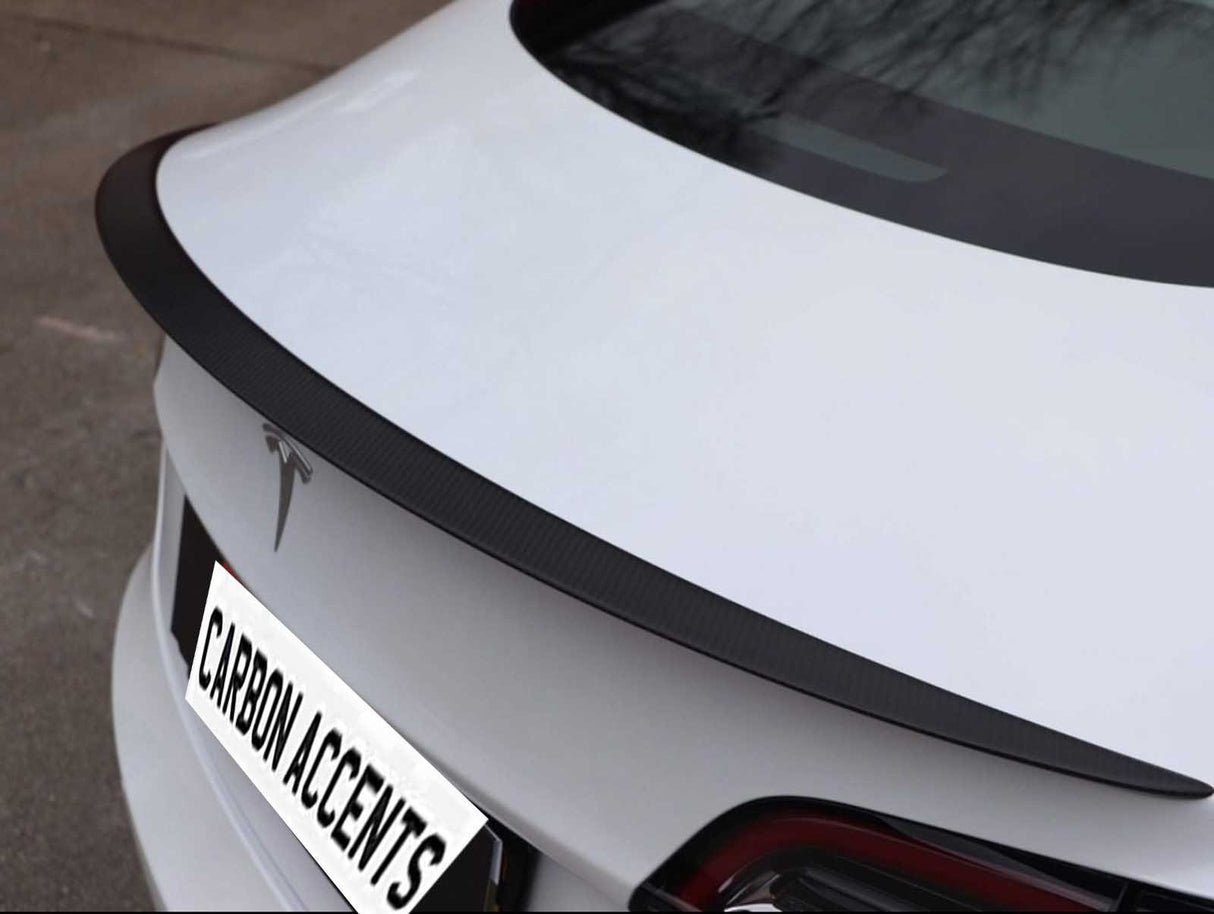 A white car features a sleek, black carbon fiber spoiler on its trunk. The car is parked outdoors, displaying a logo and a license plate reading "CARBON ACCENTS."