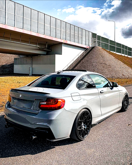 A gray BMW M235i is parked on a paved road beside a grassy mound and under a bridge, with a cloudy sky above.
