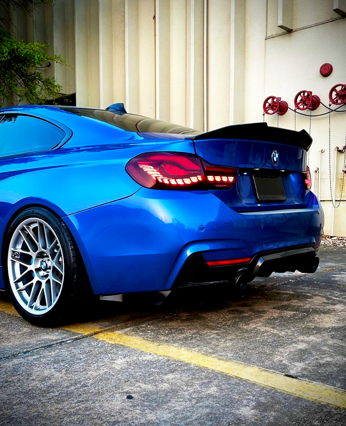 A blue sports car is parked on concrete, with tail lights illuminated. Red industrial valves are mounted on a beige wall in the background.