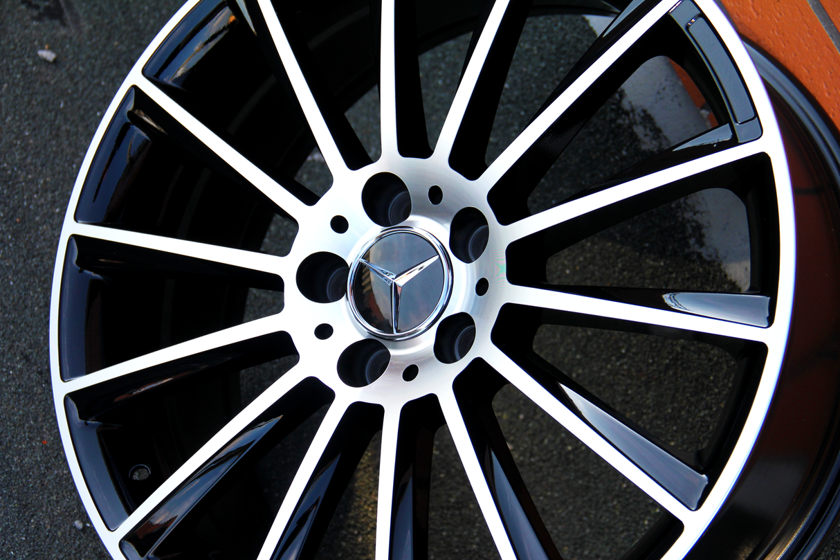 Metallic black and silver car wheel rim with spokes, featuring a three-pointed star logo at the center. The rim rests on a flat, textured surface.