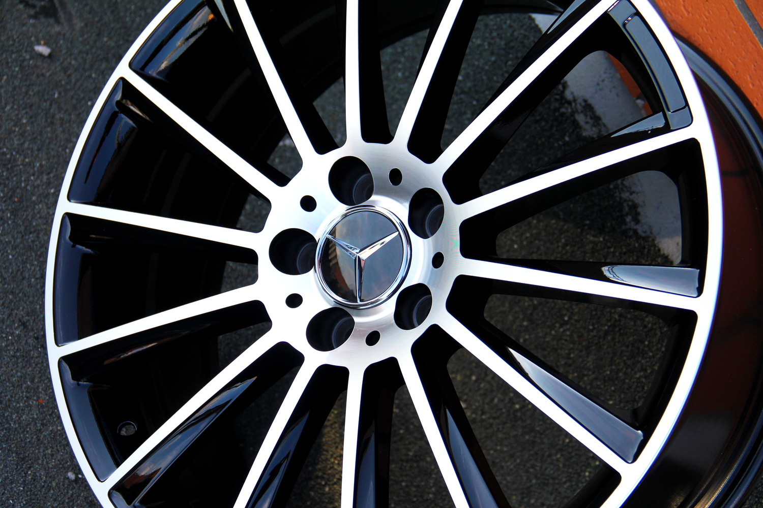 Metallic black and silver car wheel rim with spokes, featuring a three-pointed star logo at the center. The rim rests on a flat, textured surface.