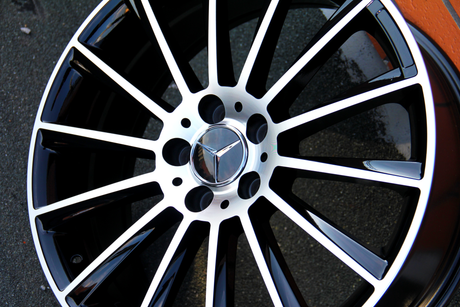 Metallic black and silver car wheel rim with spokes, featuring a three-pointed star logo at the center. The rim rests on a flat, textured surface.