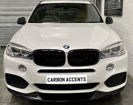 A white BMW SUV is parked indoors, facing forward with a sign on the front reading "CARBON ACCENTS." Surrounding are a tiled floor and walls with metal shuttering.