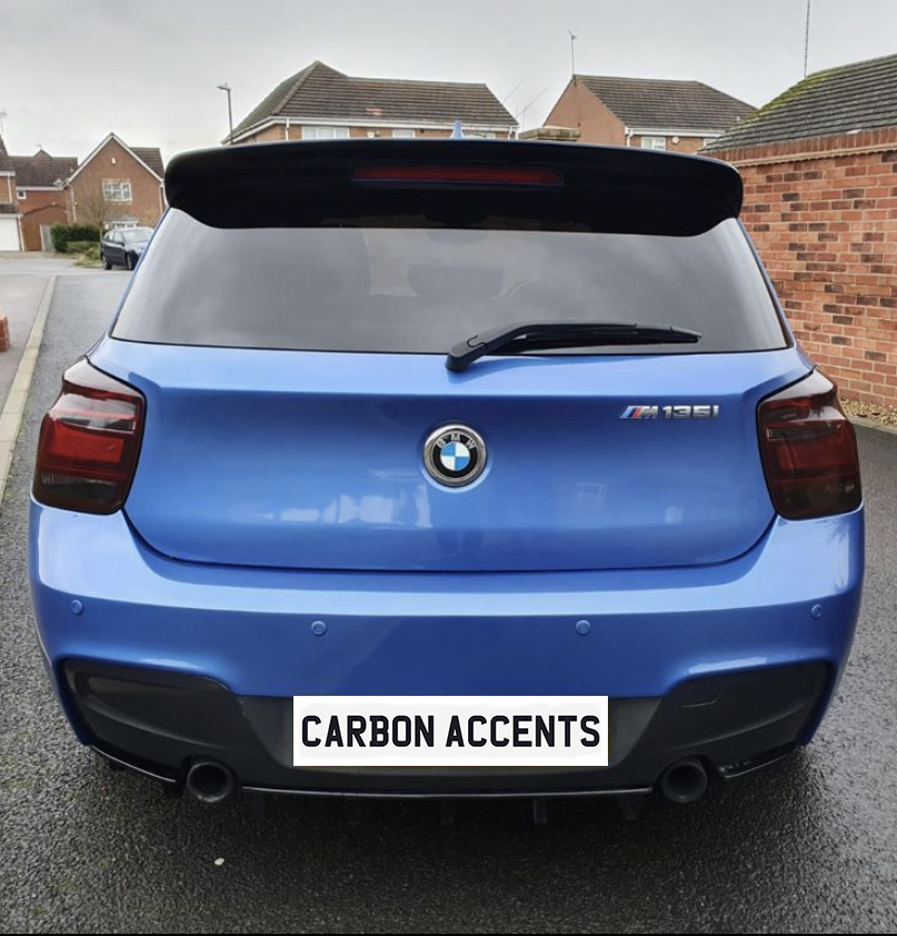 A blue BMW M135i is parked on a residential street, showing a rear view with "CARBON ACCENTS" on its bumper. Brick houses and a driveway provide the background.
