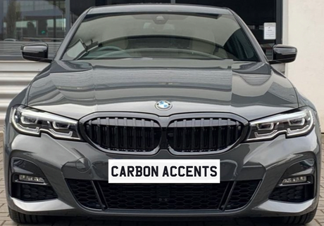A gray BMW car is parked, facing forward with sleek design elements. Text on the front reads "CARBON ACCENTS." It's positioned outdoors on a brick surface.