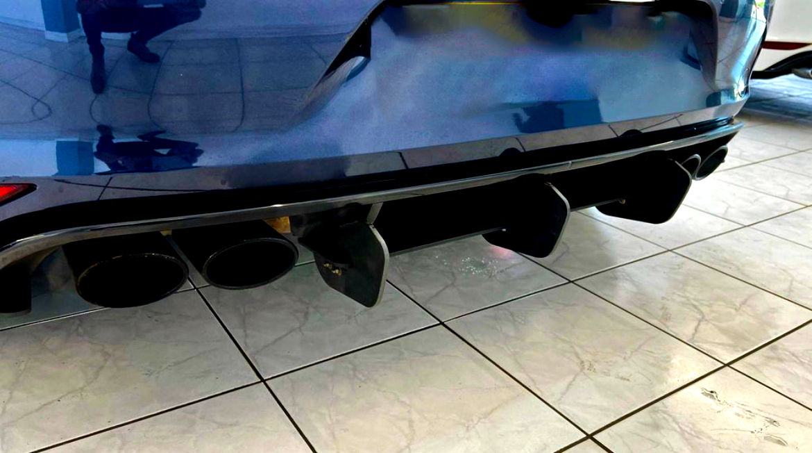 Four black exhaust pipes are protruding from the rear of a blue car, positioned over glossy tiled flooring in an indoor showroom setting.