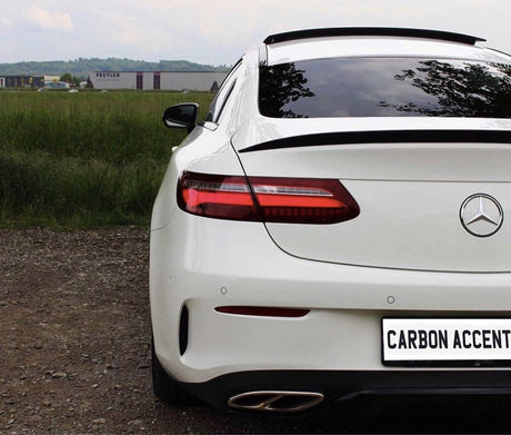 A white Mercedes-Benz car is parked on gravel, facing away. The license plate reads "CARBON ACCENT." In the background, a grassy field and industrial buildings are visible.