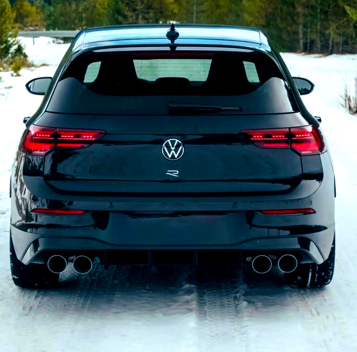 A black Volkswagen car is parked on a snow-covered road in a forested area, showing dual exhaust pipes and illuminated tail lights.