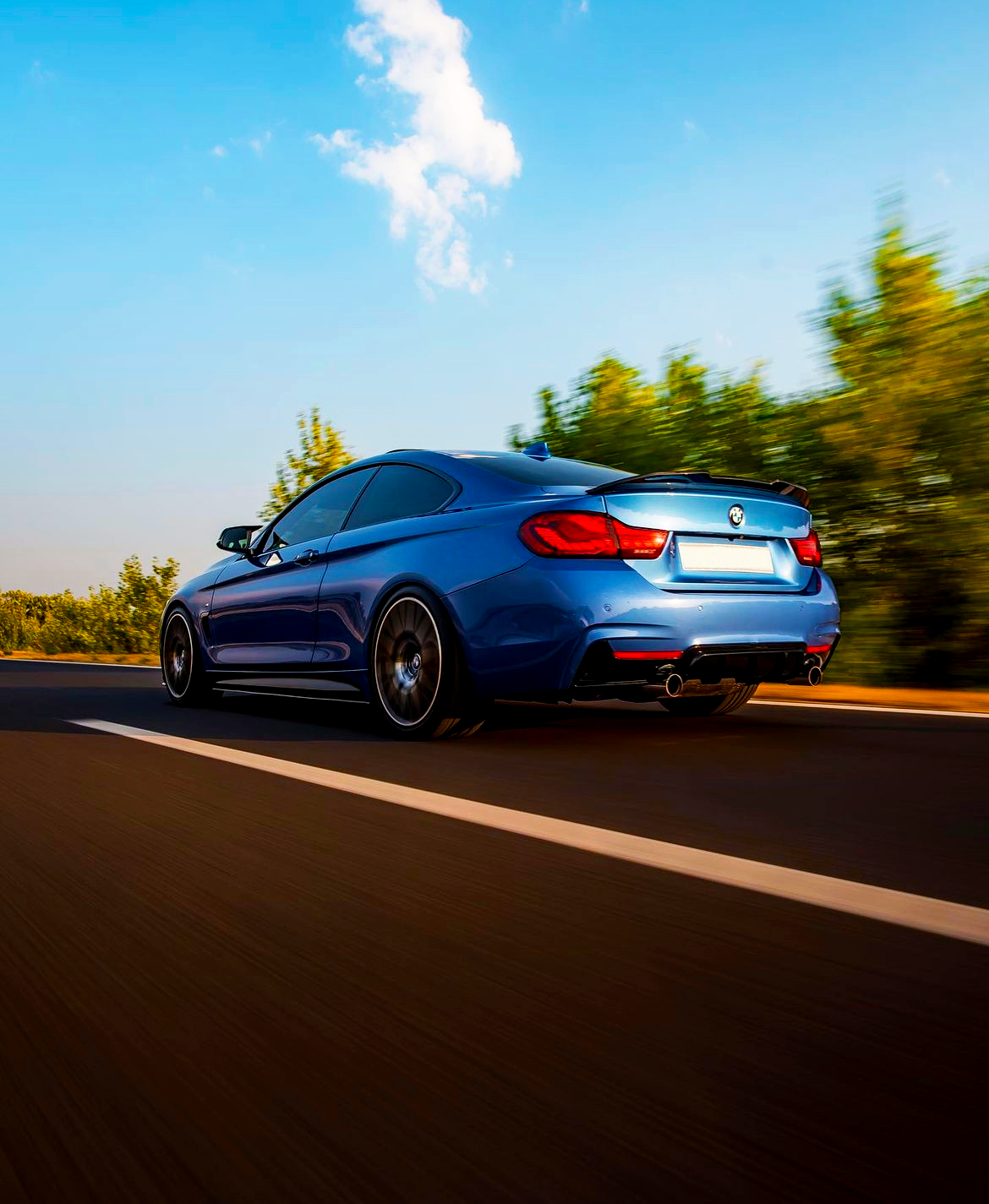 A blue sports car speeds along a two-lane highway with trees blurred in the background, under a clear blue sky with a few clouds.