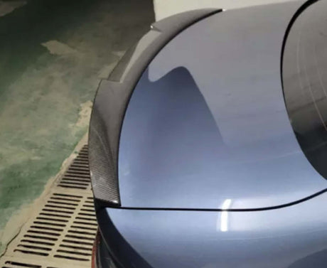 A car trunk with a carbon fiber spoiler is parked in an indoor garage with a green and beige floor, next to a drainage grate.