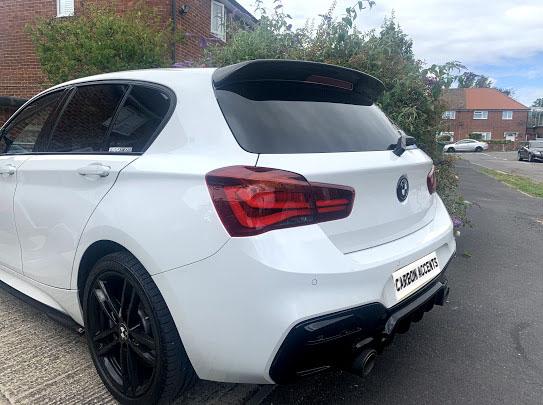 A white car with tinted windows and a rear spoiler is parked on a residential street. The license plate reads “CARBON ACCENTS”; nearby are brick houses and greenery.