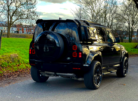 A black SUV is parked on a quiet road, with a grassy area and leafless trees surrounding it. Text "DEFENDER" is visible on the spare tire cover.