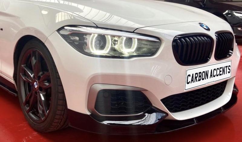 A white car with illuminated headlights and a prominent grille is on a red surface. The license plate reads "CARBON ACCENTS." Other cars are partially visible in the background.
