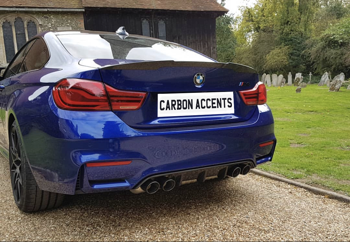 A blue BMW with the license plate "CARBON ACCENTS" is parked on a gravel path near a historic church building, surrounded by lush greenery and a small graveyard.