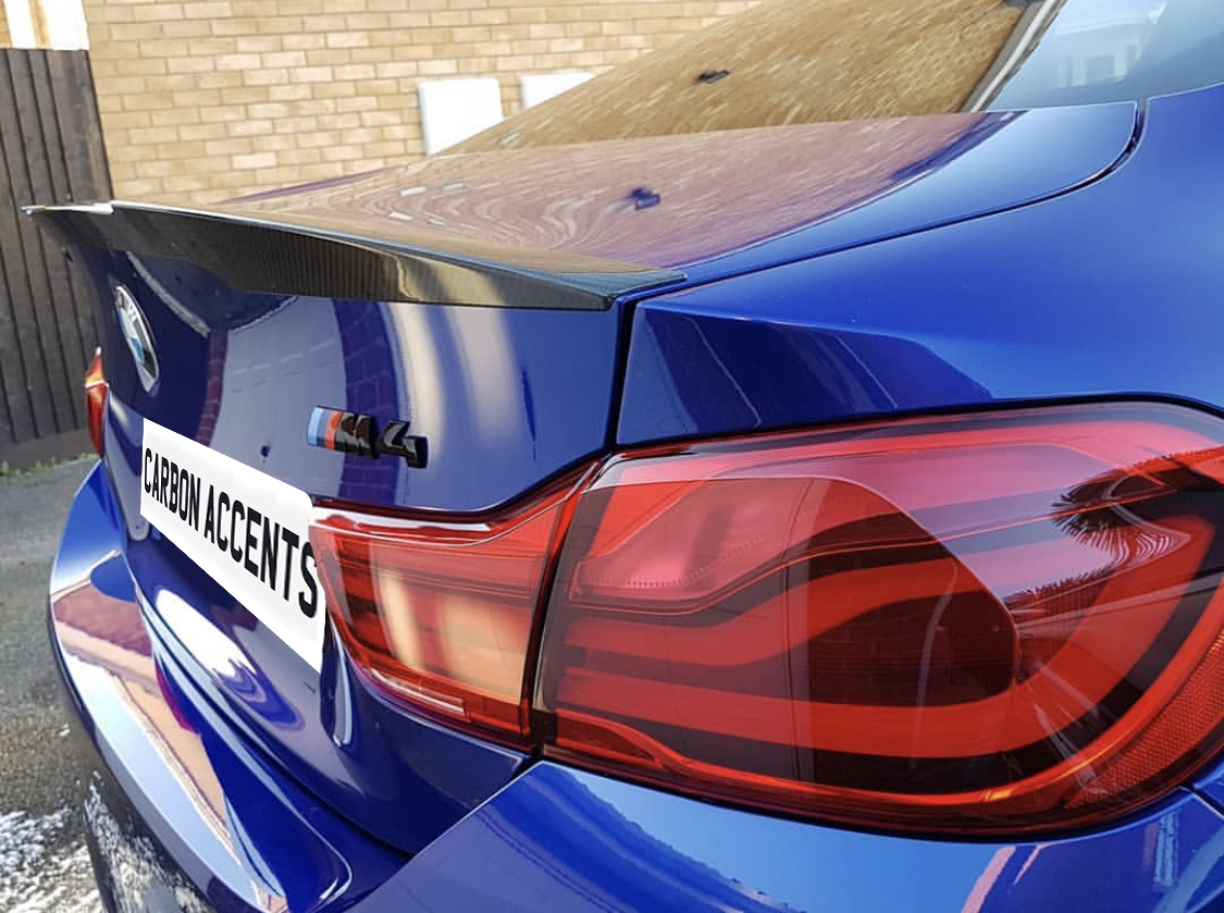A blue BMW car with a carbon fiber spoiler is parked near a brick wall. The license plate reads "CARBON ACCENTS," and there's an "M4" badge visible.