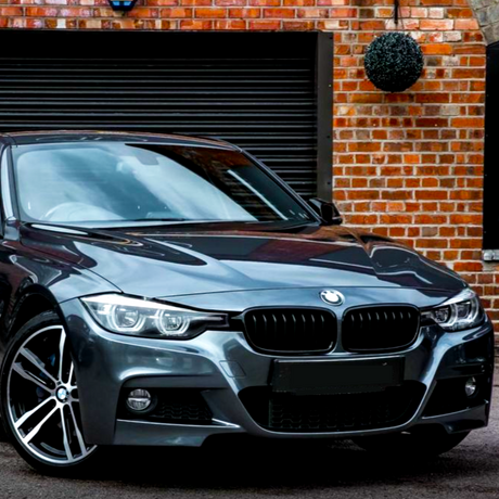 A sleek, metallic gray car is parked on a paved surface, in front of a brick wall with a hanging plant and a closed, black garage door.