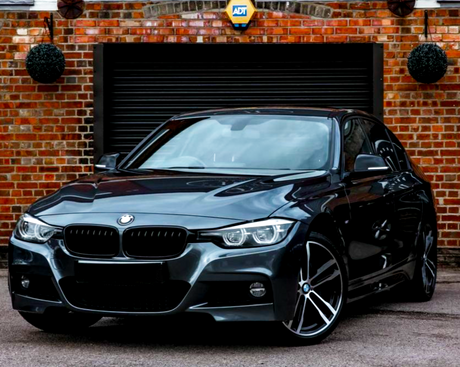 A gray BMW car is parked on a driveway in front of a brick garage with a black door. The ADT security sign is fixed above the door.