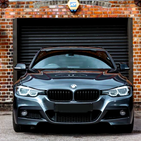 A sleek black car is parked facing forward in front of a closed black garage door. The surrounding brick wall features an "ADT" security sign.