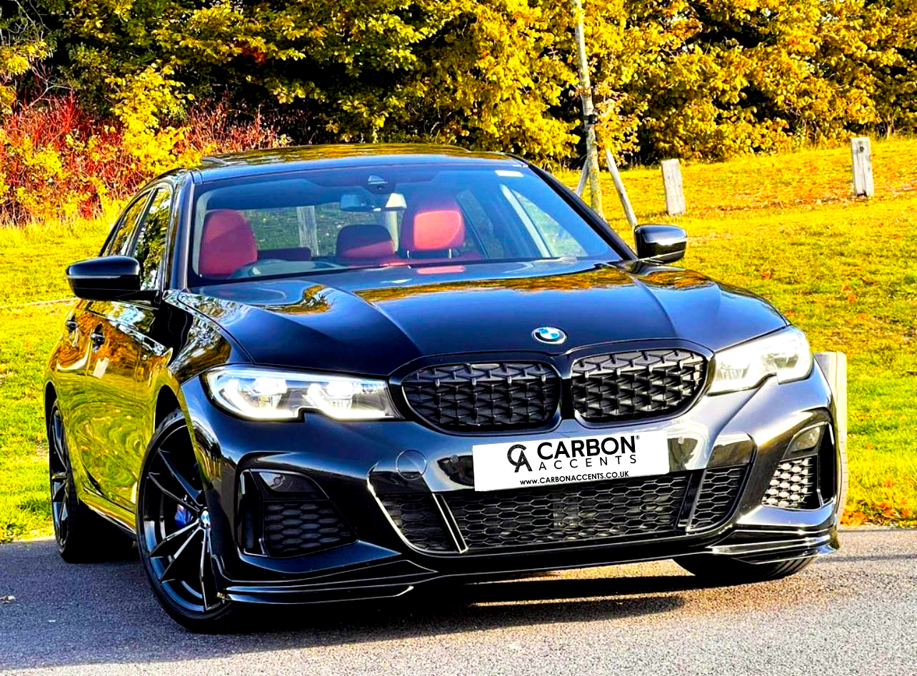 A black car with red seats is parked on a paved surface. Behind it, trees display autumn colors. The license plate reads: "CARBON ACCENTS www.CARBONACCENTS.CO.UK".