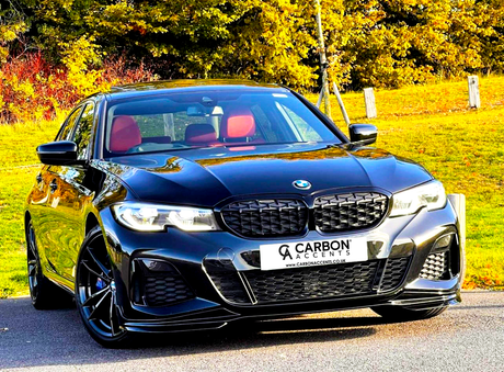 A black car with red seats is parked on a paved surface. Behind it, trees display autumn colors. The license plate reads: "CARBON ACCENTS www.CARBONACCENTS.CO.UK".