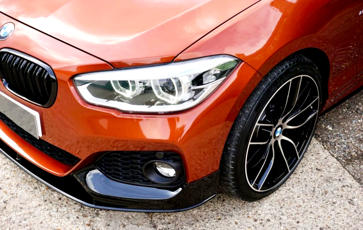 A red car with black rims is parked, displaying its BMW emblem, sleek headlights, and shiny front bumper on a textured asphalt and gravel surface.