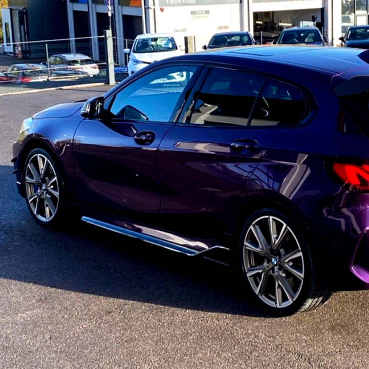 A parked purple hatchback with shiny metallic wheels sits on a paved lot, surrounded by other parked cars and a commercial building in the background.