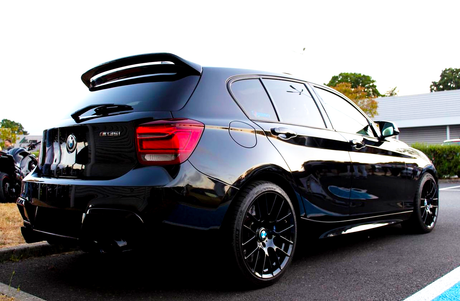 A black BMW 135i parked in a lot, featuring tinted windows, a rear spoiler, and glossy finish. Surrounding context includes greenery and a building.