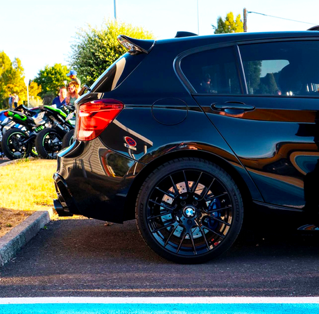 A black car with custom rims is parked beside a blue line in a lot. In the background, several motorcycles are parked near green bushes and a pole.